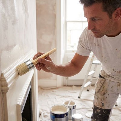 Homme peignant une cheminée blanche avec un pinceau large, pots de peinture au sol 