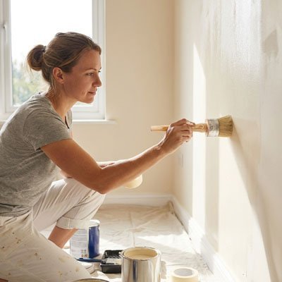 Femme peignant un mur blanc à genoux, pinceau rond en main, pots de peinture au sol, lumière naturelle