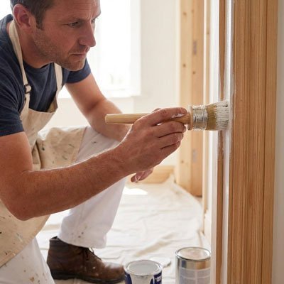 Homme peignant un encadrement de boiserie en bois clair avec un pinceau rond, tablier de travail, pots de peinture au sol 