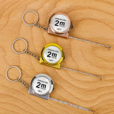 Ruban mètre porte-clés 2m acier inoxydable – 3 coloris bronze, or et argent sur fond bois, ruban déroulé