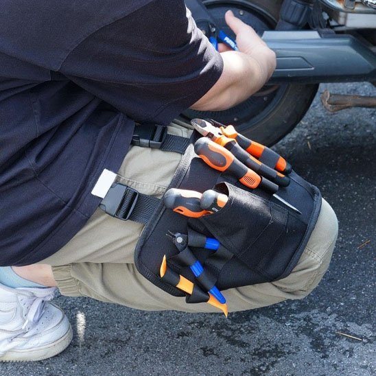 Artisan accroupi portant un sac à outils pour taille noir rempli de pinces et tournevis à manches orange et bleu