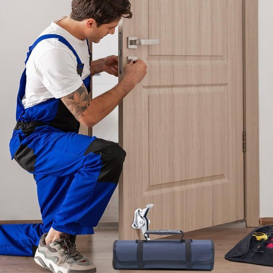 Artisan en salopette bleue installant une serrure de porte avec un sac à outils enroulable gris posé au sol à côté de lui