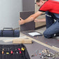 Artisan en t-shirt rouge installant des meubles de cuisine avec un sac à outils enroulable bleu et un sac noir déroulé montrant les outils organisés au premier plan