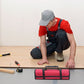 Artisan en salopette noire et t-shirt rouge accroupi avec un sac à outils enroulable rouge et noir posé devant lui sur un chantier