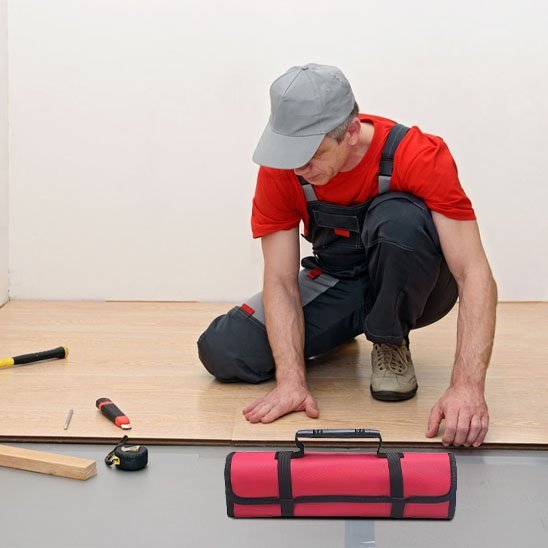 Artisan en salopette noire et t-shirt rouge accroupi avec un sac à outils enroulable rouge et noir posé devant lui sur un chantier