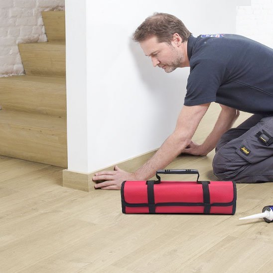 Artisan en t-shirt noir installant du parquet avec un sac à outils enroulable rouge et noir posé à côté de lui